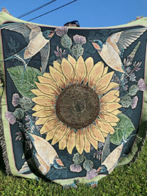 Photograph of a 72" square woven cotton throw with an image of a large sunflower surrounded by swallows and thistle plants