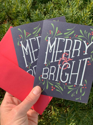 Holiday greeting card with the hand-lettered slogan Merry & Bright surrounded by mistletoe leaves and berries