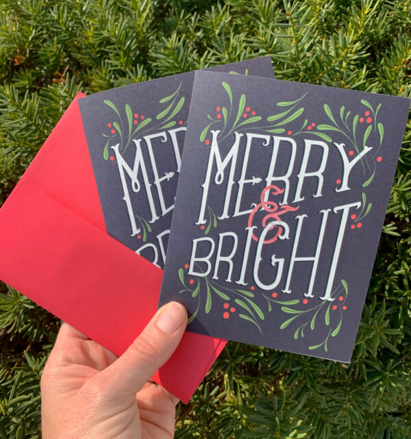 Holiday greeting card with the hand-lettered slogan Merry & Bright surrounded by mistletoe leaves and berries