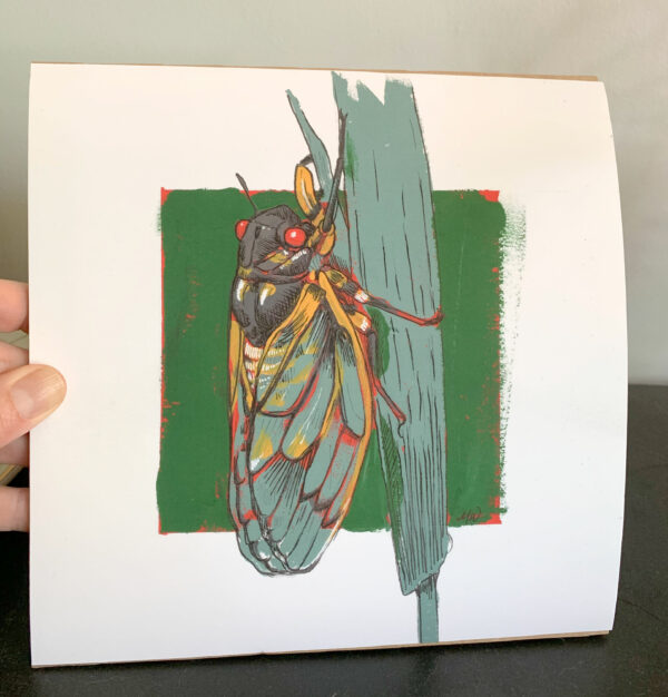 A hand holding an 8x8 art print of a cicada painted in gouache